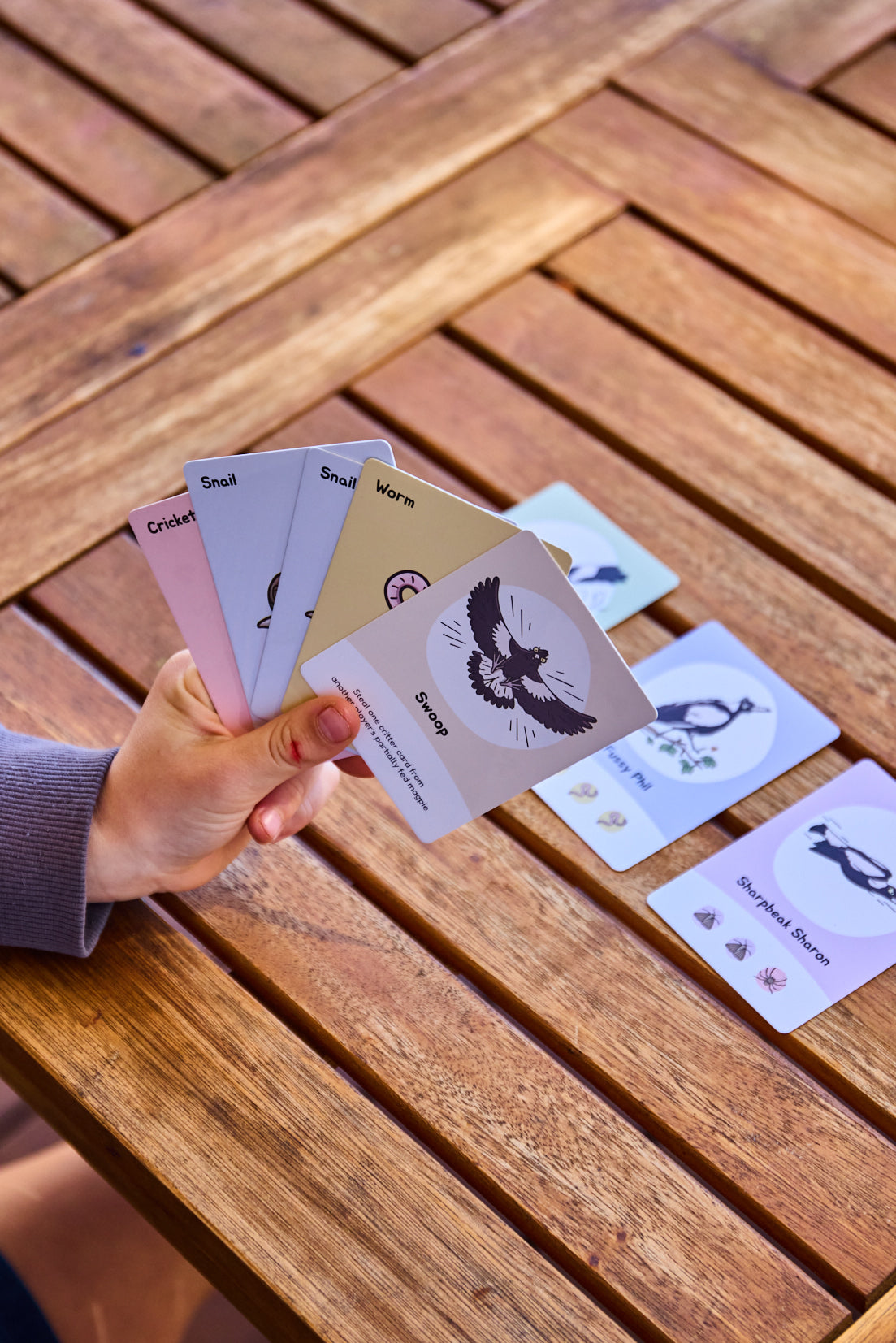 A hand holds a fan of Swoopy Season cards over a wooden slatted table, with several cards laid out on the table.