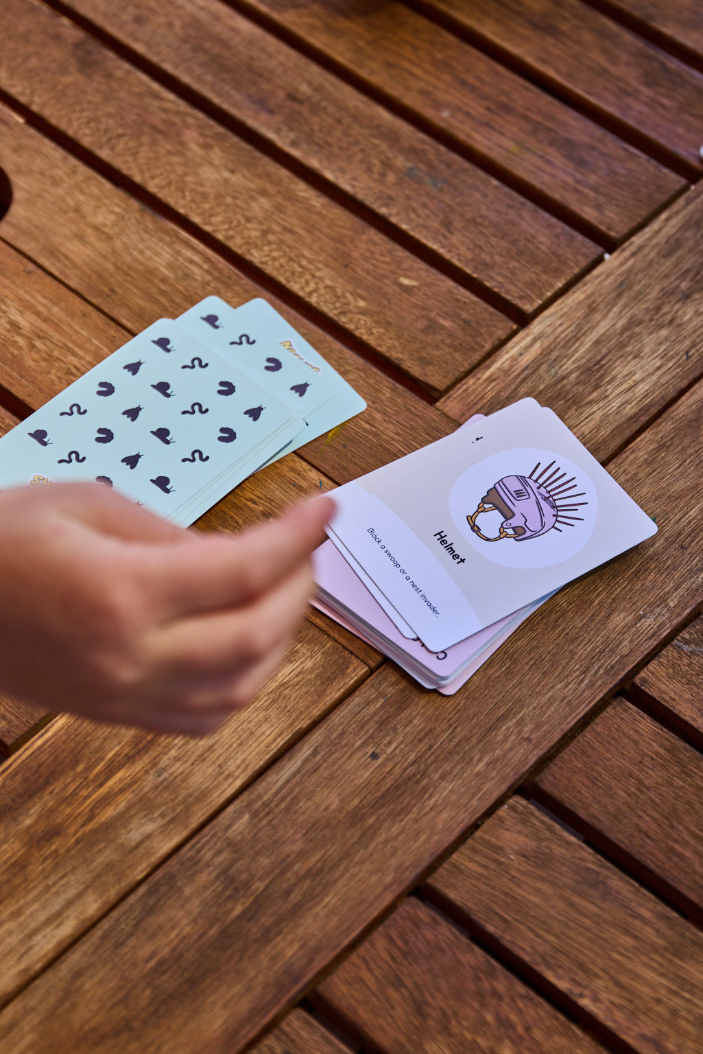 A hand is placing cards from Swoopy Season on a brown wooden slatted table. One card is face up, showing a purple illustration of a character with the word 'Helmet' printed below it, and another card is fanned out behind it showing light blue backs with black symbols.
