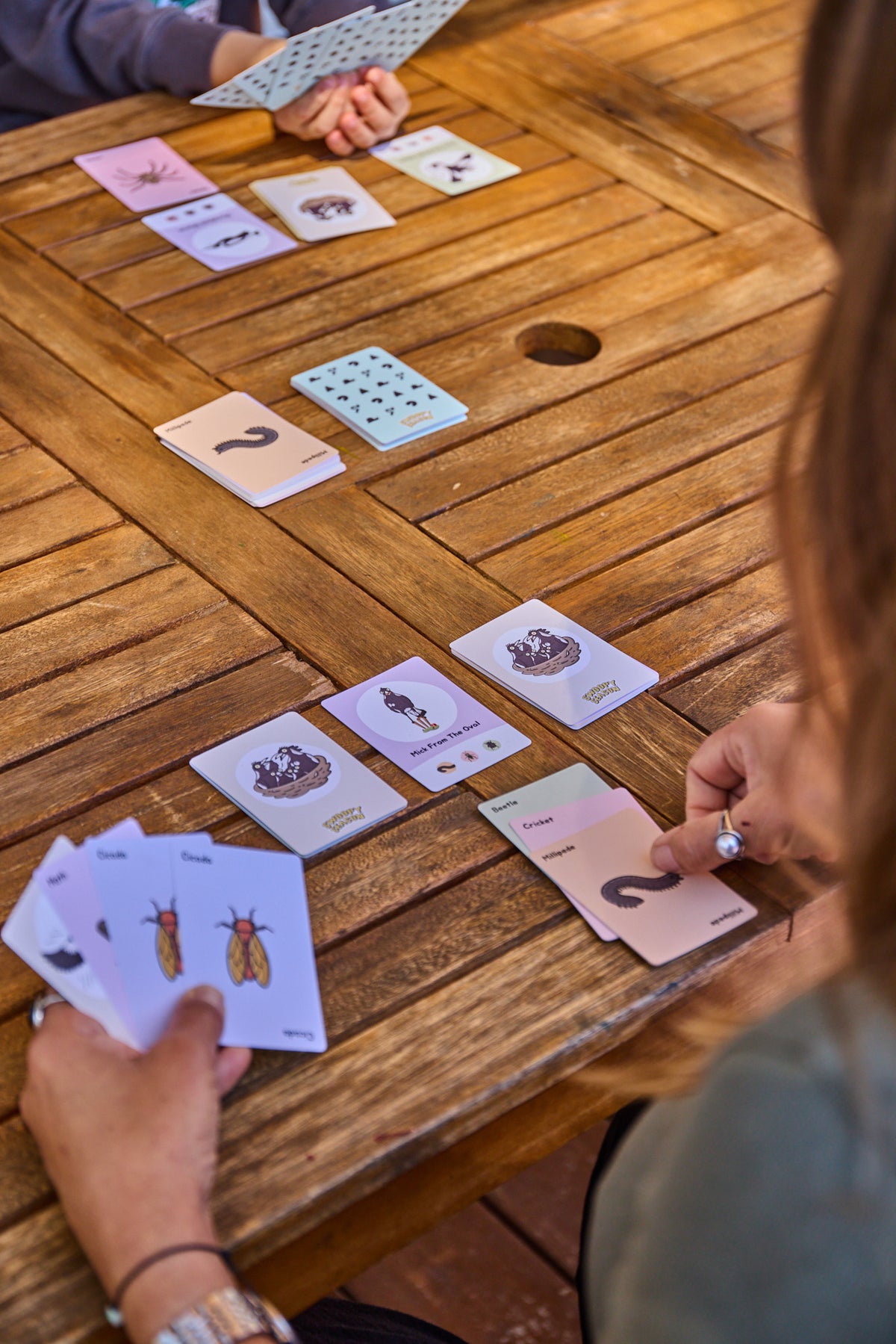 A close-up, over-the-shoulder view of people playing a card game at a wooden outdoor table. Several hands are visible holding and turning pastel-colored cards with illustrations and symbols, while more cards are laid out on the table.