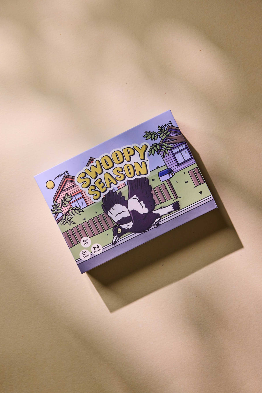 A square product box featuring an illustration of a magpie, with the title 'SWOOPY SEASON' in large yellow letters, photographed on a beige surface with soft shadow overlays.