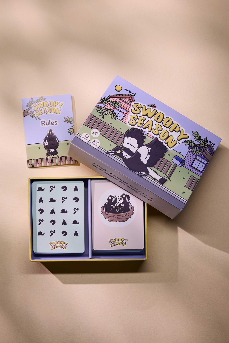 A boxed card game set titled 'Swoopy Season' arranged on a warm beige surface. The set includes a decorative outer box, a smaller card sheet labeled 'Rules,' and an open tray of cards with illustrated card backs.