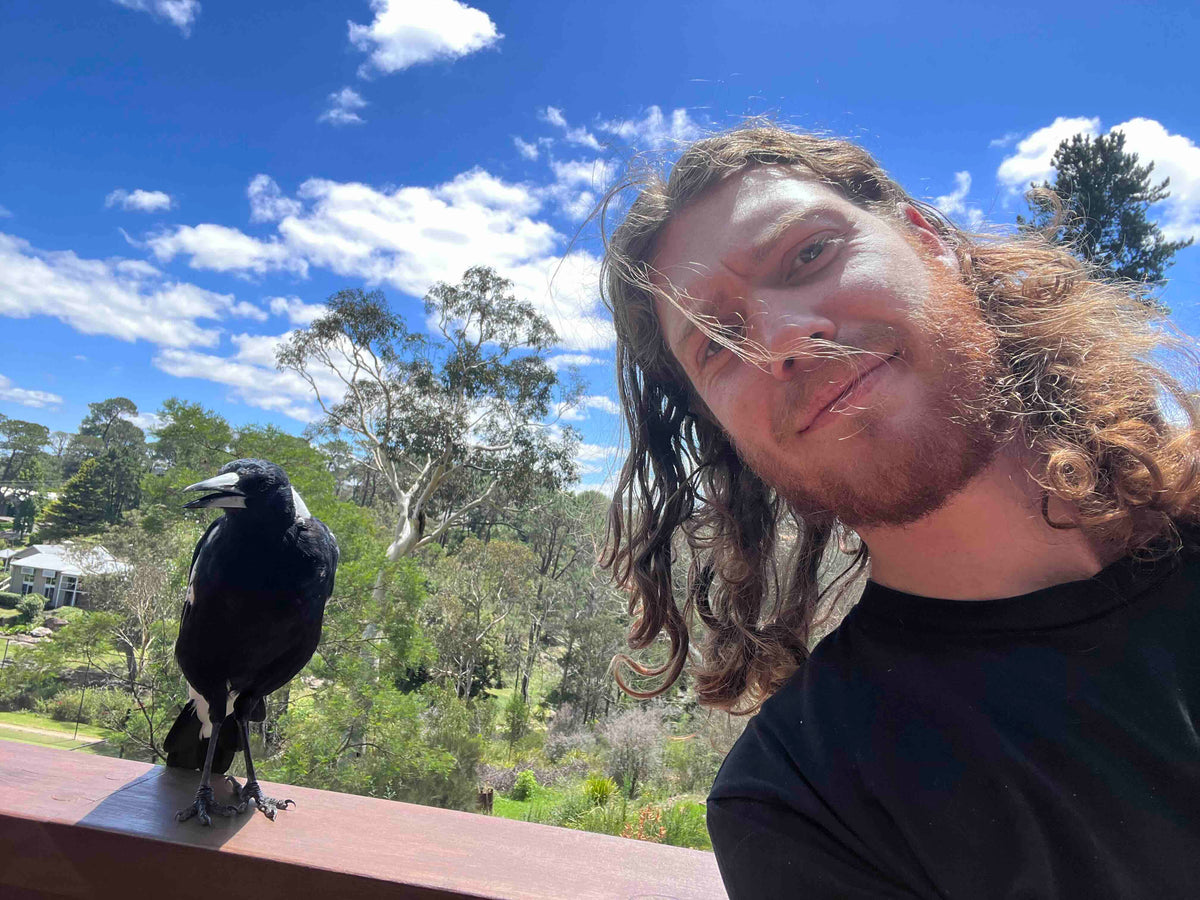 A man with long wavy light brown hair and a beard takes a selfie outdoors on a sunny day, standing next to a magpie perched on a wooden railing.