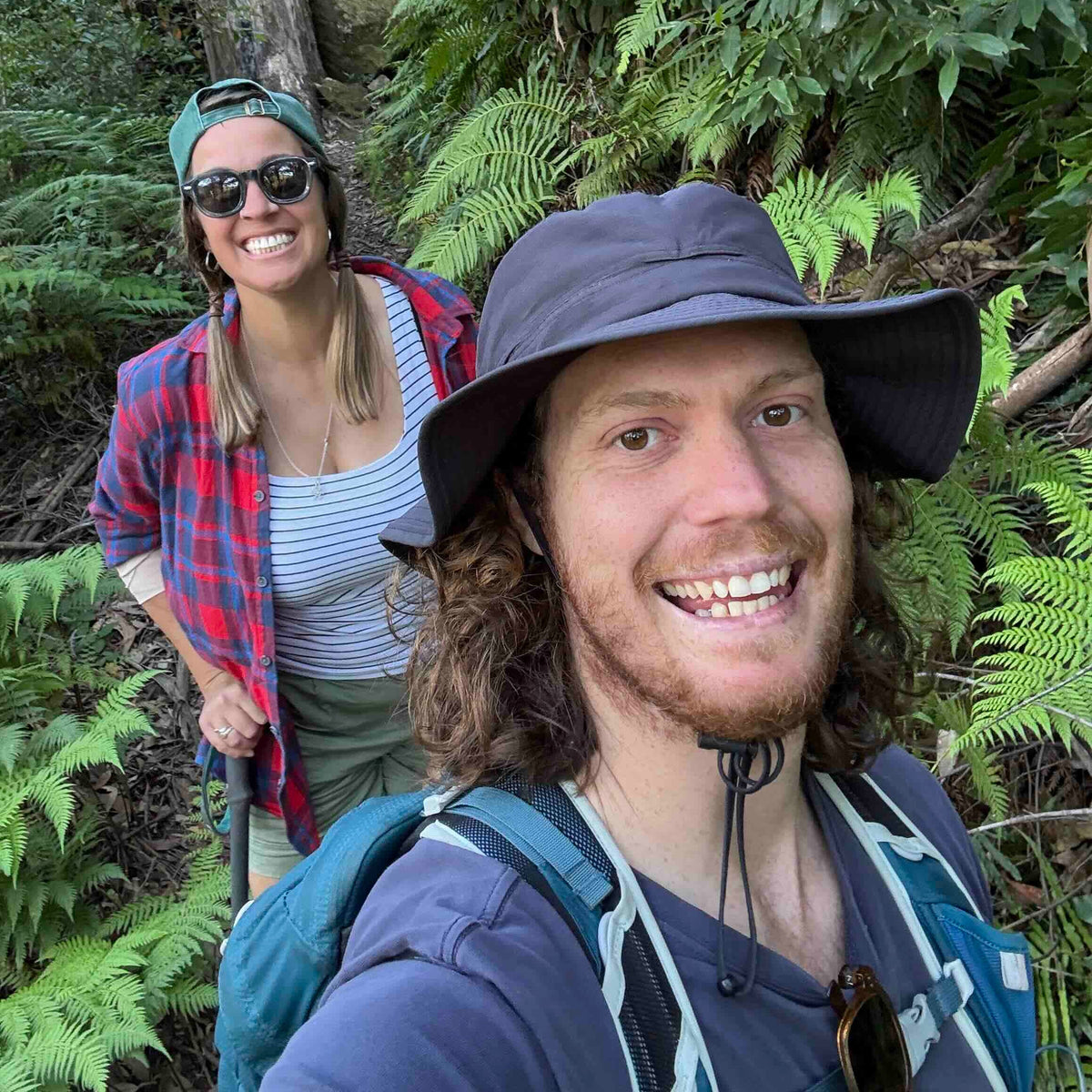 A smiling man in a wide-brimmed dark hat and blue shirt takes a selfie while a woman in sunglasses, a red plaid shirt, and a teal backpack stands behind him on a lush green forest trail.
