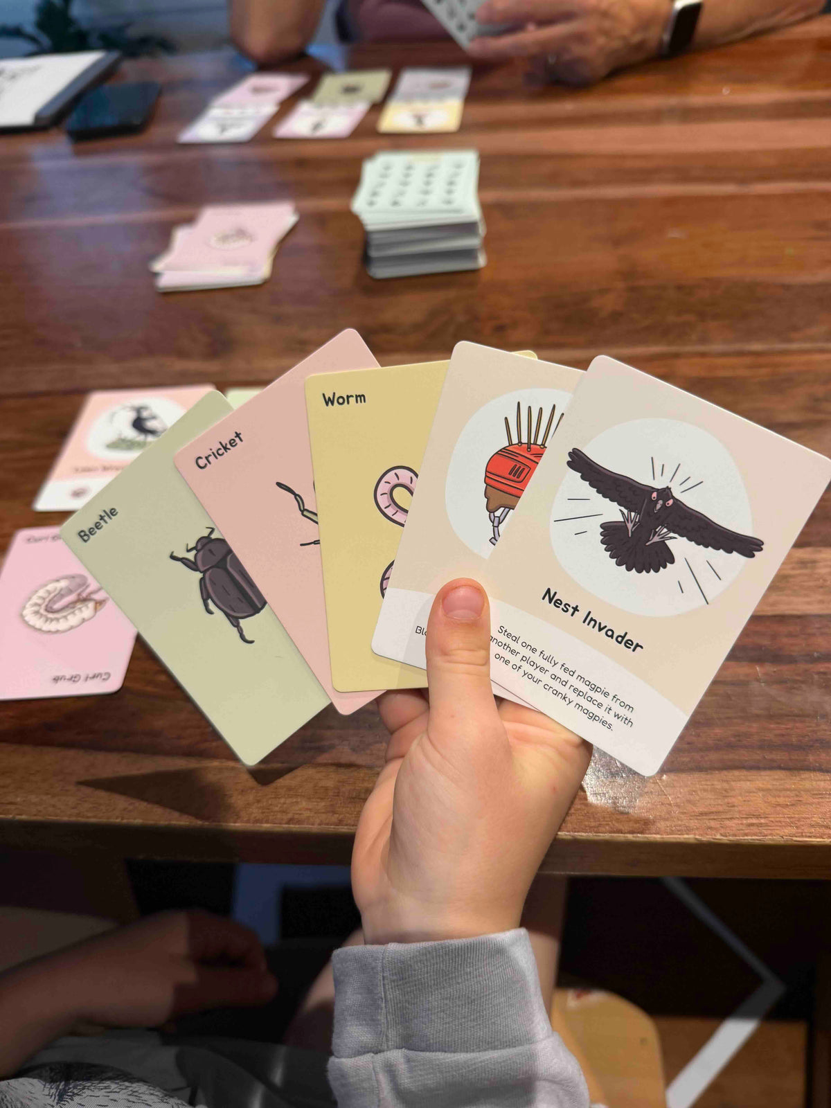 A hand holds a fan of illustrated Swoopy Season card over a wooden table, showing cards titled Beetle, Cricket, Worm, and Nest Invader.